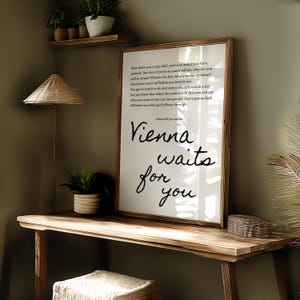 May include: A framed print with the words "Vienna waits for you" in black script. The print is displayed on a wooden desk with a woven lamp and a small potted plant. The background is a muted green.