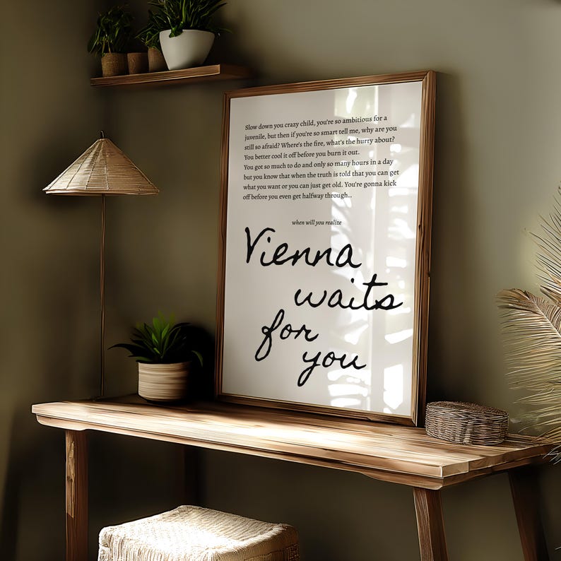 May include: A framed print with the words "Vienna waits for you" in black script. The print is displayed on a wooden desk with a woven lamp and a small potted plant. The background is a muted green.