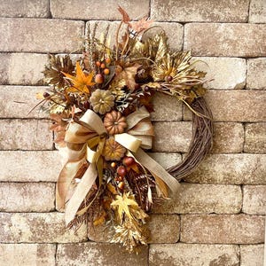 May include: A seasonal wreath with a brown grapevine base, decorated with artificial pumpkins, leaves, berries, and a large tan ribbon. The wreath has gold and orange tones, creating a warm autumnal decoration.