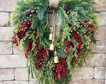 May include: A Christmas wreath featuring green foliage, red berries, and gold embellishments. The wreath includes pinecones and small gold bells, suspended by a natural rope. It is displayed against a brick wall, creating a seasonal decoration.