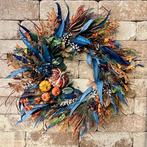 May include: A fall-themed wreath with a variety of textures and colors. It features blue, orange, and brown leaves, berries, and small pumpkins. The wreath is set against a brick wall.