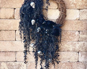 May include: A Halloween wreath with a natural grapevine base, adorned with black foliage, silver skulls, and black bird figures. The wreath is displayed against a brick wall, creating a spooky and decorative accent for the holiday season.