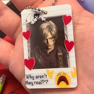 Op de afbeelding: Een rechthoekige sleutelhanger met een foto van een persoon met blond haar, omringd door rode harten. De sleutelhanger heeft de tekst "Why aren't they real??" en een huilende emoji.