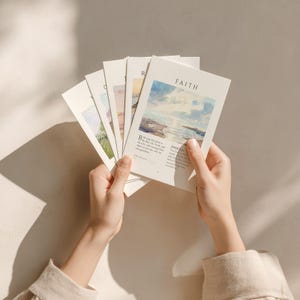 Puede incluir: Un conjunto de tarjetas rectangulares en abanico, cada una con una ilustración de paisaje estilo acuarela y texto impreso. La tarjeta superior muestra la palabra FAITH sobre una escena costera y un pasaje de Juan 3:16.