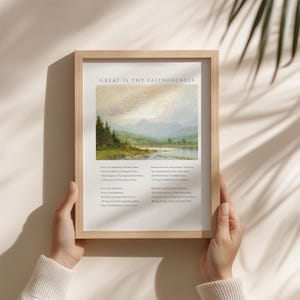 May include: A framed print with the text "GREAT IS THY FAITHFULNESS" above a landscape painting of a lake and mountains. The print is held by hands and has a light wood frame. The painting features trees, water, and a cloudy sky.
