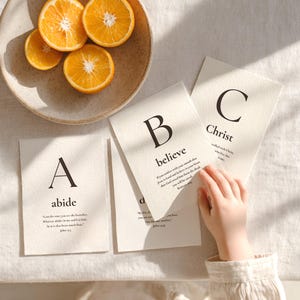May include: Three off-white rectangular cards featuring black serif letters A, B, and C with corresponding words abide, believe, and Christ, along with Bible verses. A speckled bowl containing four orange slices sits nearby.