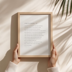 Könnte beinhalten: Gerahmter Druck mit dem Text von Sprüche 31. Der Text ist in schwarzer Schrift auf cremefarbenem Hintergrund. Der Rahmen ist hellbraun. Der Druck wird von zwei Händen gehalten.