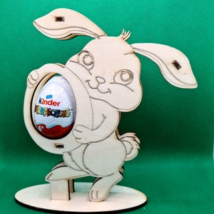 May include: Wooden bunny rabbit figurine holding a Kinder Surprise egg. The bunny is light brown and has a white face with black eyes. The egg is red and white with the words "Kinder Surprise" on it.