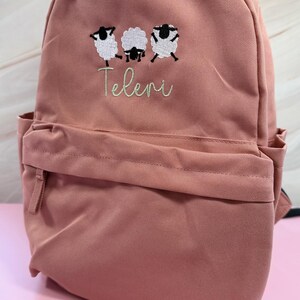 May include: A dusky pink backpack with a front pocket and two shoulder straps. The backpack has an embroidered design of three sheep and the word "Teleri" in a cursive font.