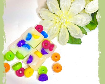 May include: A white background with colorful wax melts shaped like fruit loops. The wax melts are arranged in a square shape with a white flower in the background. The text "Fruit Loops" is written in red and yellow letters at the bottom of the image.