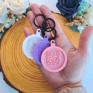 Puede incluir: Tres llaveros redondos en blanco, morado y rosa, cada uno con un diseño de código QR. Los llaveros están unidos por anillas negras. El fondo incluye una rodaja de madera y arreglos florales.