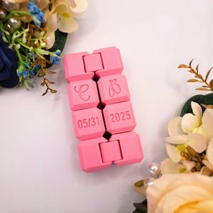 May include: A bright pink, articulated fidget toy, featuring the initials "C" and "B" and the date "05/31 2025". The toy is made of connected, cube-shaped segments. The background includes floral arrangements with white and blue flowers.
