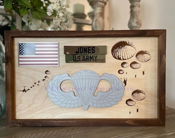 May include: A wooden shadow box with a brown frame, featuring a US flag, a US Army name tag with the name "JONES", a silver parachute wings emblem, and a laser-engraved design of parachutes and paratroopers.