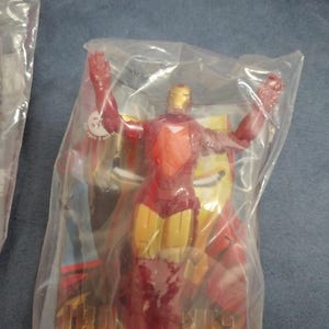May include: A red and gold Iron Man action figure in a plastic bag. The figure is posed with arms outstretched. The bag has a picture of Iron Man on it with the text "Iron Man 2".