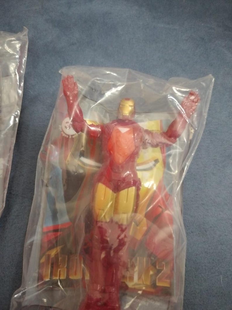 May include: A red and gold Iron Man action figure in a plastic bag. The figure is posed with arms outstretched. The bag has a picture of Iron Man on it with the text "Iron Man 2".