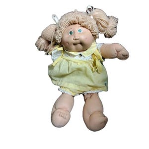以下が含まれることがあります： ブロンドの糸のツインテールと青い目を持つ、ヴィンテージのキャベツパッチキッズ人形。人形は白いレースのトリムが付いた黄色のドレスを着ています。人形の腕と脚は柔らかく、顔はバラ色の肌色をしています。