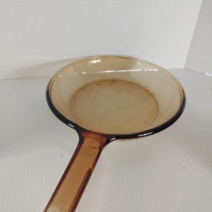 Puede incluir: Una sartén de vidrio marrón con un mango largo. La sartén está vacía y tiene una superficie texturizada.