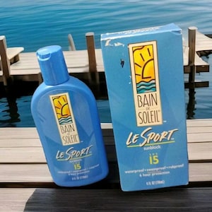May include: A blue bottle of Bain de Soleil Le Sport sunblock SPF 15 with a yellow sun logo. The bottle is sitting on a wooden surface next to a blue box with the same logo and text. The box says "waterproof, sweatproof, rubproof, 6 hour protection".