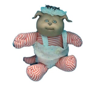 May include: A vintage plush doll with a round face, small ears, and blue eyes. The doll wears a red and white striped shirt and matching striped trousers. It has a white nappy with a floral pattern and a blue and white head covering. The doll is a collectible item.