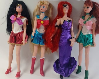 May include: Four Sailor Moon character dolls. The dolls are dressed in their signature outfits. The doll on the left is wearing a red and white outfit with a black bow. The doll in the middle is wearing a blue and white outfit with a red bow. The doll on the right is wearing a green and white outfit with a pink bow. The doll in the back is wearing a purple dress.