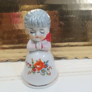 May include: A white porcelain bell shaped like a girl with closed eyes, wearing a red bow and a floral dress with orange and blue flowers.