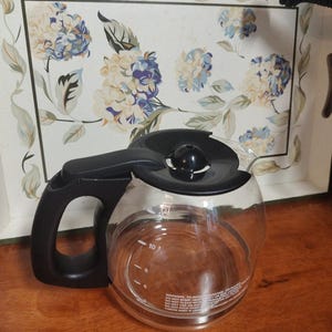 以下が含まれることがあります： 黒い蓋と取っ手のある透明なガラス製のコーヒーポット。ポットには、6、8、10杯分の目盛りがあります。ポットは木製の表面の上に置かれています。