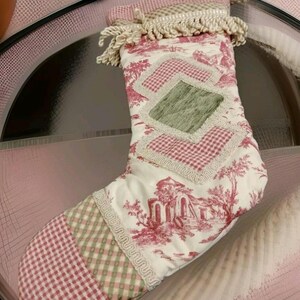 Può includere: Un calzino di Natale rosa e verde con sfondo bianco e una frangia bianca in cima. Il calzino ha un design a patchwork con un motivo a quadri verde e bianco, un motivo a quadri rosa e bianco e un motivo floreale.
