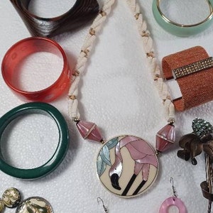 Puede incluir: Un conjunto de joyas que incluye un collar con un colgante de flamenco rosa, un par de pendientes de concha rosa con detalles de perlas y varios brazaletes en verde, marrón, rojo y beige.