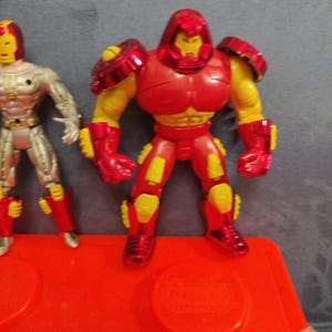 May include: Two action figures, one red and yellow and one silver and yellow, stand on a red plastic base. The red and yellow figure has a helmet and is wearing armor. The silver and yellow figure is wearing a suit.