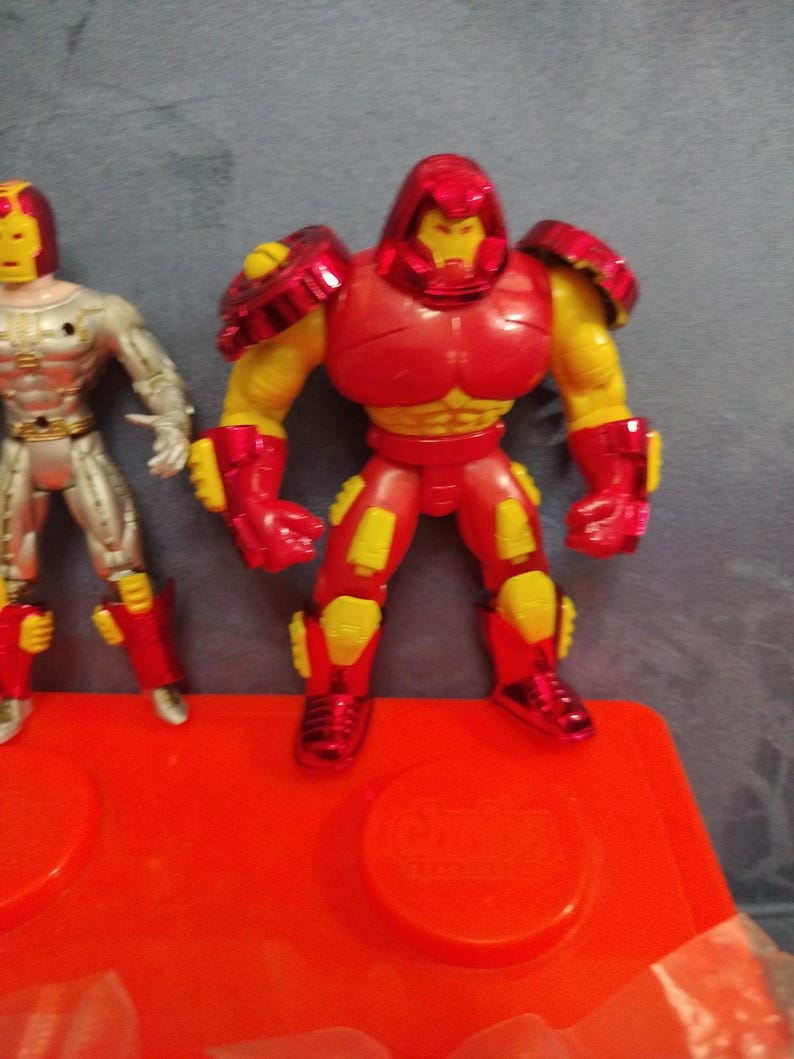 May include: Two action figures, one red and yellow and one silver and yellow, stand on a red plastic base. The red and yellow figure has a helmet and is wearing armor. The silver and yellow figure is wearing a suit.