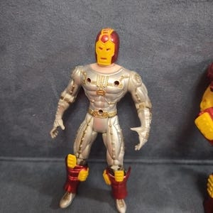 May include: Silver and red action figure of a superhero wearing a helmet with a yellow face mask. The figure has a muscular build and is posed in a standing position.