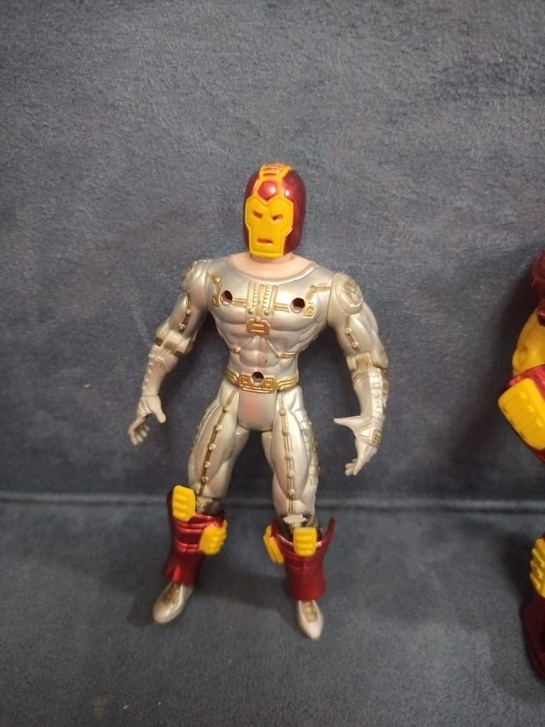 May include: Silver and red action figure of a superhero wearing a helmet with a yellow face mask. The figure has a muscular build and is posed in a standing position.