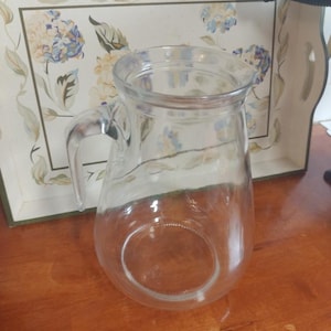 Puede incluir: Una jarra de vidrio transparente con asa, colocada sobre una superficie de madera. La jarra está vacía y tiene una base redondeada. Una bandeja decorativa con motivos florales se encuentra en el fondo.