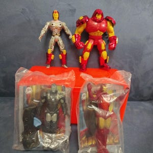 May include: Four Iron Man action figures in plastic packaging. The figures are red, silver, and black. The figures are from the Marvel Comics universe.