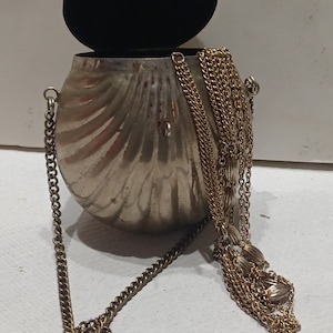 May include: A silver-colored, shell-shaped handbag with a hinged lid and a black interior. The bag features a long, gold-colored chain strap and a decorative gold chain tassel. The clasp is gold-colored.