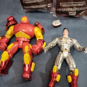 May include: Two action figures, one red and one silver, with yellow accents. The red figure is Iron Man, and the silver figure is a Marvel character. Both figures are posed in action stances.
