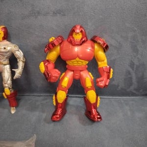 May include: A red and yellow action figure of a superhero wearing a helmet and armor. The figure is standing on a gray surface.
