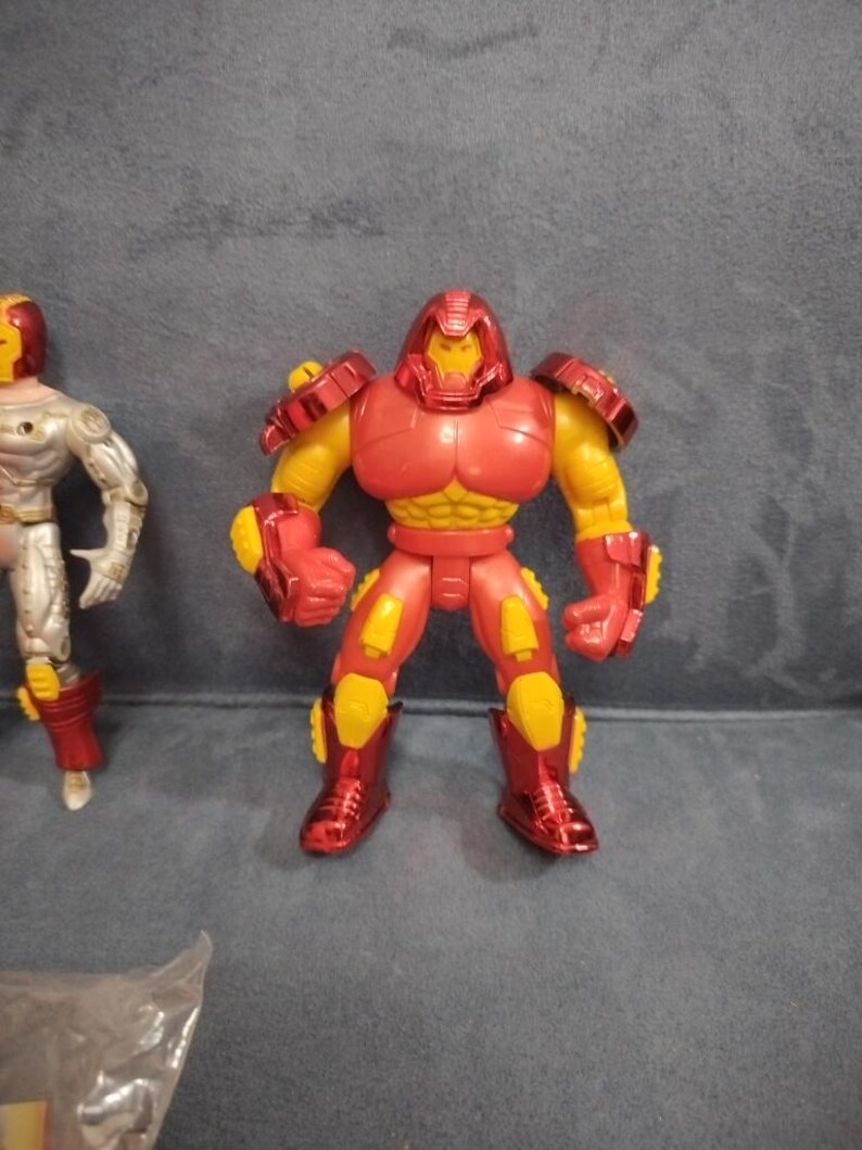 May include: A red and yellow action figure of a superhero wearing a helmet and armor. The figure is standing on a gray surface.