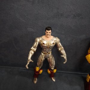 May include: Silver and gold action figure with a black mustache and hair. The figure is wearing red and yellow armor.