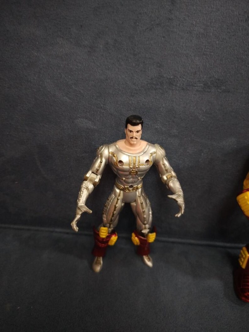May include: Silver and gold action figure with a black mustache and hair. The figure is wearing red and yellow armor.