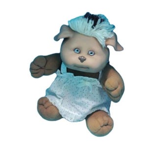 May include: A vintage plush doll with a tan face, brown arms and legs, and blue eyes. The doll has white hair with a dark brown patch and wears a white dress with a polka dot pattern. The doll's arms are raised, and it appears to be sitting.