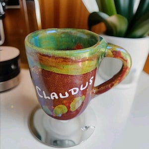 Könnte beinhalten: Eine handgefertigte Keramiktasse mit einem einzigartigen Design. Die Tasse hat einen braunen Boden mit dem Namen "CLAUDUS" in weißen Buchstaben. Der Rand ist grün und blau, mit einem gelben Streifen. Sie steht auf einem kleinen, silbernen Ständer. Eine kleine, dekorative Kaffeetasse.