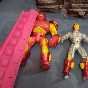 May include: Two action figures, one red and one silver, with yellow accents. The red figure is a large, muscular character with a helmet. The silver figure is a more slender character with a helmet and a silver chest plate. Both figures are standing on a gray surface. The text "MARVEL" is visible in the background.