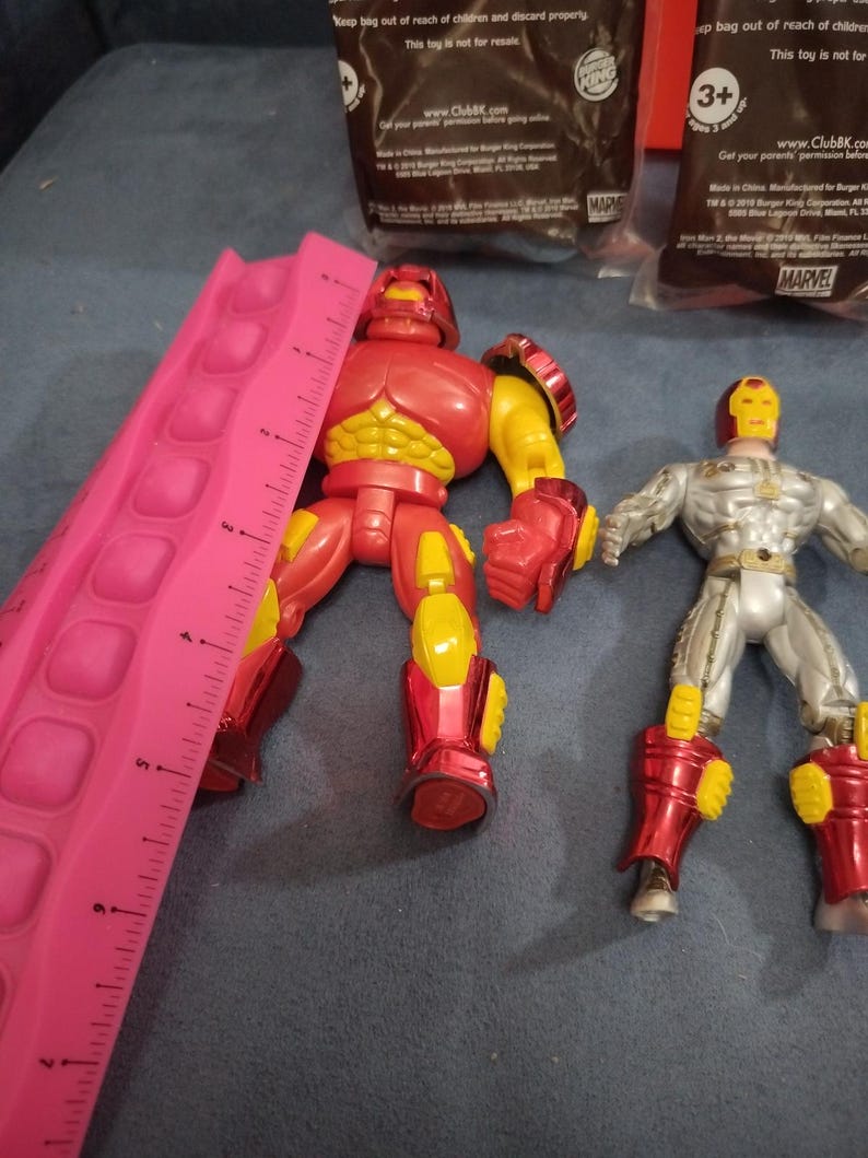 May include: Two action figures, one red and one silver, with yellow accents. The red figure is a large, muscular character with a helmet. The silver figure is a more slender character with a helmet and a silver chest plate. Both figures are standing on a gray surface. The text "MARVEL" is visible in the background.