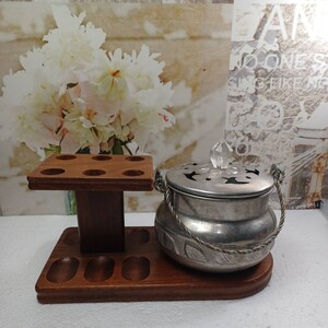 May include: A wooden pipe rack with six slots for pipes and a round, silver, decorative pot with a lid and a crystal knob. The pot has a rope handle and an ornate design.