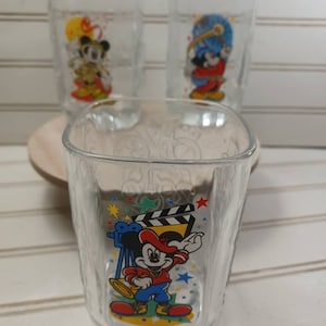 Puede incluir: Juego de tres vasos transparentes con Mickey Mouse en varias poses. El vaso delantero tiene a Mickey con un tema de cine con sombrero rojo y pantalón azul. Los otros vasos tienen diferentes diseños de Mickey Mouse.