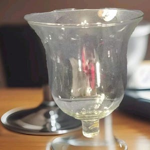 May include: A clear glass candle holder with a flared top and a rounded base. The candle holder is sitting on a wooden surface. The glass has a slightly textured appearance.