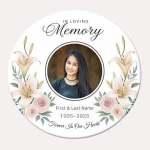 May include: A white round memorial with a floral design featuring lilies and roses. A photo of a person is in the center of the circle. The text "In Loving Memory" is at the top of the circle. The text "First & Last Name" is below the photo. The text "1995-2025" is below the name. The text "Forever In Our Hearts" is at the bottom of the circle.