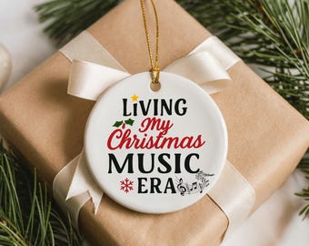 May include: A white ceramic Christmas ornament with the text "Living My Christmas Music Era" in black, red, and green. The ornament is hanging on a gold string and is placed on a wrapped gift with a cream ribbon.