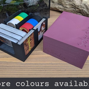 May include: A black organiser for the board game Splendor, with compartments for cards and coloured chips. The organiser is next to a purple box with the word "Splendor" printed on it.
